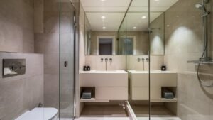 A beautifully designed small bathroom showcasing smart space-saving strategies. The room features a corner sink, wall-mounted faucet, and floating vanity, all contributing to a streamlined and modern look. Large-scale tiles on the floor and walls, along with a glass shower door, create a spacious feel, while light colors and a large mirror enhance brightness and the perception of space. The overall aesthetic is clean, minimalistic, and functional.